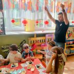 Mężczyzna stojący na środku pomieszczeni z uniesionymi rękoma w których trzyma bambusowe pałeczki do jedzenia sushi. Przy stoliku siedzą dzieci ubrane w białe fartuszki i opaski na czołach. Przed nimi na stoliku znajdują się talerze z porcją sushi. Dzieci uczą się posługiwać pałeczkami.