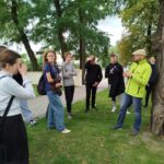 Prowadzący warsztaty questingu rozmawia z grupą uczniów przy dużym drzewie przed kościołem.