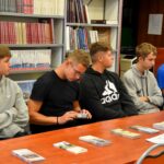 Uczniowie siedzą przy stole w bibliotece podczas warsztatów questingu, na stole leżą ulotki i materiały informacyjne.
