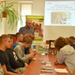 Prowadzący warsztaty questingu przemawia do uczniów w bibliotece, w tle widoczna jest prezentacja na ekranie.