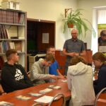 Prowadzący warsztaty questingu stoi przed uczniami siedzącymi przy stole w bibliotece, na stole leżą ulotki i materiały.