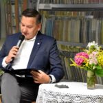 Zbigniew Czerkas - wicestarosta Powiatu Sokołowskiego siedzi przy stole na którym stoi kolorowy bukiet kwiatów i czyta wiersze Jana Kochanowskiego.