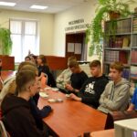Uczniowie siedzą przy długim stole w bibliotece podczas warsztatów questingu, na stole leżą materiały i ulotki.