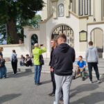 Uczniowie i prowadzący warsztaty stoją i siedzą przed bramą kościoła w Sokołowie Podlaskim podczas zajęć terenowych questingu.
