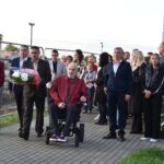 Kilkanaście osób stoi tuż przed złożeniem kwiatów pod pomnikiem.