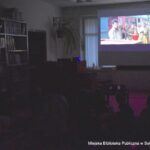 Czytelnia w bibliotece. Scena w mroku. Na ścianie wyświetlany jest film dla dzieci.