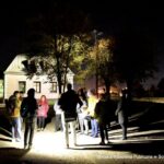 Nocna scena na zewnątrz, obok biblioteki podczas spaceru ulicą Kolejową. Wokół pracowników pracowni dokumentacji dziejów miasta stoi kilka osób i słucha przemówienia jednego z pracowników.