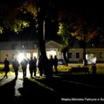 Nocna scena na zewnątrz, przed biblioteką podczas spaceru ulicą Kolejową. Wokół pracowników pracowni dokumentacji dziejów miasta stoi kilka osób i słucha przemówienia jednego z pracowników.