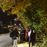 Nocna scena na zewnątrz, na chodniku przy ulicy Kolejowej. podczas spaceru ulicą Kolejową. Wokół pracowników pracowni dokumentacji dziejów miasta stoi kilka osób i słucha przemówienia jednego z pracowników.