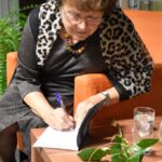 Renata Kopczewska podpisuje książkę. Siedzi na fotelu, obok na stoliku leży pomarańczowa róża.