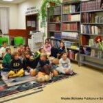 Sala czytelni w bibliotece. Na dywanie siedzą dzieci podczas Nocy bibliotek. Przed nimi stoi bibliotekarka, która prowadzi konkurs dla dzieci.