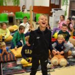Sala czytelni w bibliotece. Na dywanie siedzą dzieci podczas Nocy bibliotek. Na pierwszym planie stoi chłopiec i odpowiada na pytania.