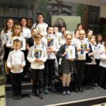 Wszystkie dzieci, które brały udział w konkursie recytatorskim, w każdej z trzech kategorii wiekowych. Dzieci trzymają w rękach dyplomy oraz nagrody książkowe. Obok stoi komisja oraz dyrektor biblioteki Małgorzata Iwańska-Kania.