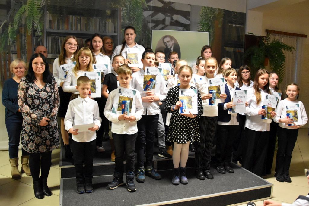 Wszystkie dzieci, które brały udział w konkursie recytatorskim, w każdej z trzech kategorii wiekowych. Dzieci trzymają w rękach dyplomy oraz nagrody książkowe. Obok stoi komisja oraz dyrektor biblioteki Małgorzata Iwańska-Kania.