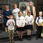 Czwórka nagrodzonych dzieci w pierwszej grupie wiekowej. Dzieci trzymają w rękach nagrody książkowe oraz dyplomy. Za nimi trzyosobowa komisja oraz dyrektor biblioteki Małgorzata Iwańska-Kania.