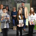 Trójka nagrodzonych dzieci w drugiej grupie wiekowej. Dzieci trzymają w rękach nagrody książkowe oraz dyplomy. Za nimi trzyosobowa komisja oraz dyrektor biblioteki Małgorzata Iwańska-Kania.