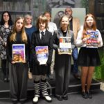 Czwórka nagrodzonych dzieci w trzeciej grupie wiekowej. Dzieci trzymają w rękach nagrody książkowe oraz dyplomy. Za nimi trzyosobowa komisja oraz dyrektor biblioteki Małgorzata Iwańska-Kania.