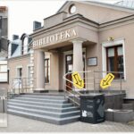 Zdjęcie przed budynkiem biblioteki. Dwie żółte strzałki wskazują miejsce, gdzie znajduje się wyrzutnia. Jest po lewej stronie przed wejściem na schody. Na zdjęciu umieczone są też dwa inne zdjęcia pokazujące samo urządzenie - wrzutnię, z boku i od przodu.