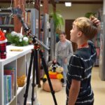 Chłopiec na pierwszym planie spogląda w ekran telefonu zamontowanego na statywie, podczas pracy nad projektem warsztatowym.