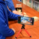 Smartfon na statywie. W tle widać ręce dzieci, które ustawiają drewniane klocki w czytelni.