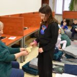 „Dziecko odbiera kartę podczas zajęć w bibliotece, w tle inni uczestnicy.