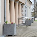 Przed wejściem z tyłu budynku do Miejskiej Biblioteki Publicznej stoją duże betonowe donice z roślinami tworzącymi Kolejowy Ogród Deszczowy. Obok wejścia widoczna jest pochylnia z metalową poręczą, a w tle chodnik, drzewa i ulica.