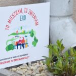 Tabliczka informująca o dofinansowaniu projektu w konkursie „Tu mieszkam, tu zmieniam EKO”, ustawiona w donicy ogrodu deszczowego obok rury spustowej. Wokół rosną młode zielone rośliny i leżą jasne kamienie.