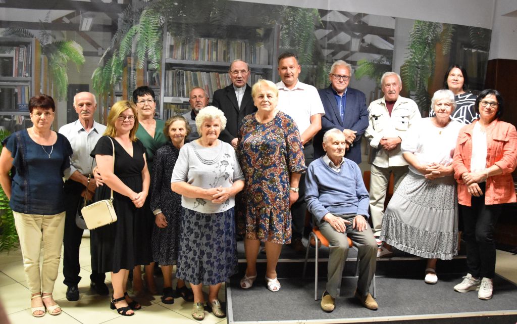 Na zdjęciu widać grupę szesnastu osób pozujących do zdjęcia w Bibliotece. Grupa składa się z osób stojących i jednej siedzącej na krześle w pierwszym rzędzie. Na zdjęciu znajdują się, sponsorzy książki Wiesławy Kwiek, przedstawiciele władz miasta i powiatu - burmistrz miasta Sokołów Podlaski Iwona Kublik oraz wicestarosta powiatu Sokołowskiego Zbigniew Czerkas oraz osoby, które opowiedziały historie do książki. W tle znajduje się baner zajmujący całą ścianę, na którym znajduje się zdjęcie pomieszczenia działu dla dorosłych z widocznymi regałami z książkami oraz kwiatami paproci na nich.