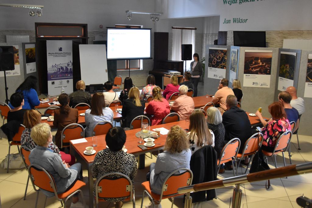 Fotografia przedstawia osoby siedzące przy stolikach, są to pracownicy biblioteka z terenu powiatu sokołowskiego. Przed nimi stoi kobieta. Przed kobietą znajduje się duży ekran do wyświetlania prezentacji.