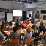 Fotografia przedstawia osoby siedzące przy stolikach, są to pracownicy biblioteka z terenu powiatu sokołowskiego. Przed nimi stoi kobieta. Przed kobietą znajduje się duży ekran do wyświetlania prezentacji.