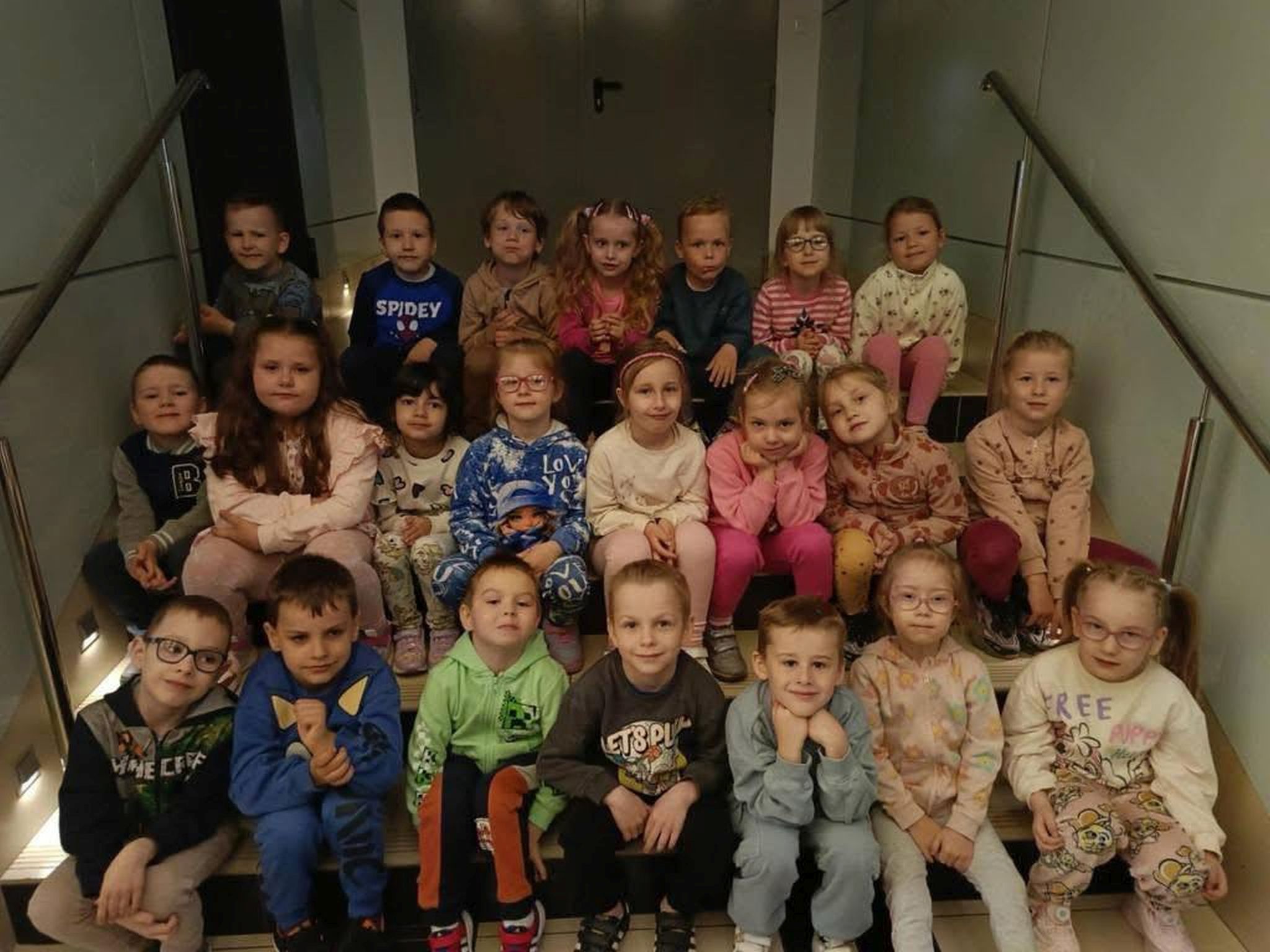 Grupa 22 dzieci (przedszkolaków) siedzi na schodach w trzech rzędach. Za nim szare dwuskrzydłowe drzwi. Dzieci znajdują się w Sokołowskim Ośrodku Kultury.