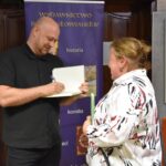 Bartłomiej Dejnega podczas podpisywania książek. Obok stoi kobieta.