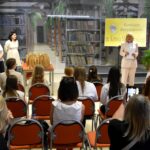 Przewodnicząca komisji konkurswe w jasnym garniturze stoi na holu biblioteki z kartkami w rękach. Obok niej za stołem z kilkunastoma papierowymi torebkami–nagrodami stoją trzy kobiety, a przed nimi na pomarańczowych krzesłach siedzą dzieci i dorośli. Z prawej widoczny żółty baner „Konkurs Recytatorski im. Kornela Makuszyńskiego”.