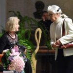 Lucyna Aleksandrowicz-Pędich siedzi na pomarańczowym fotelu, trzyma skórzane etui i patrzy w stronę stojącej przy niej starszej kobiety w białym żakiecie z brązową torebką na ramieniu.