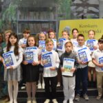Grupa dzieci trzyma w rękach nagrody książkowe oraz dyplomy. Obok po prawej i lewej stronie stoją cztery kobiety. Trzy z komisji konkursowej oraz dyrektor biblioteki.