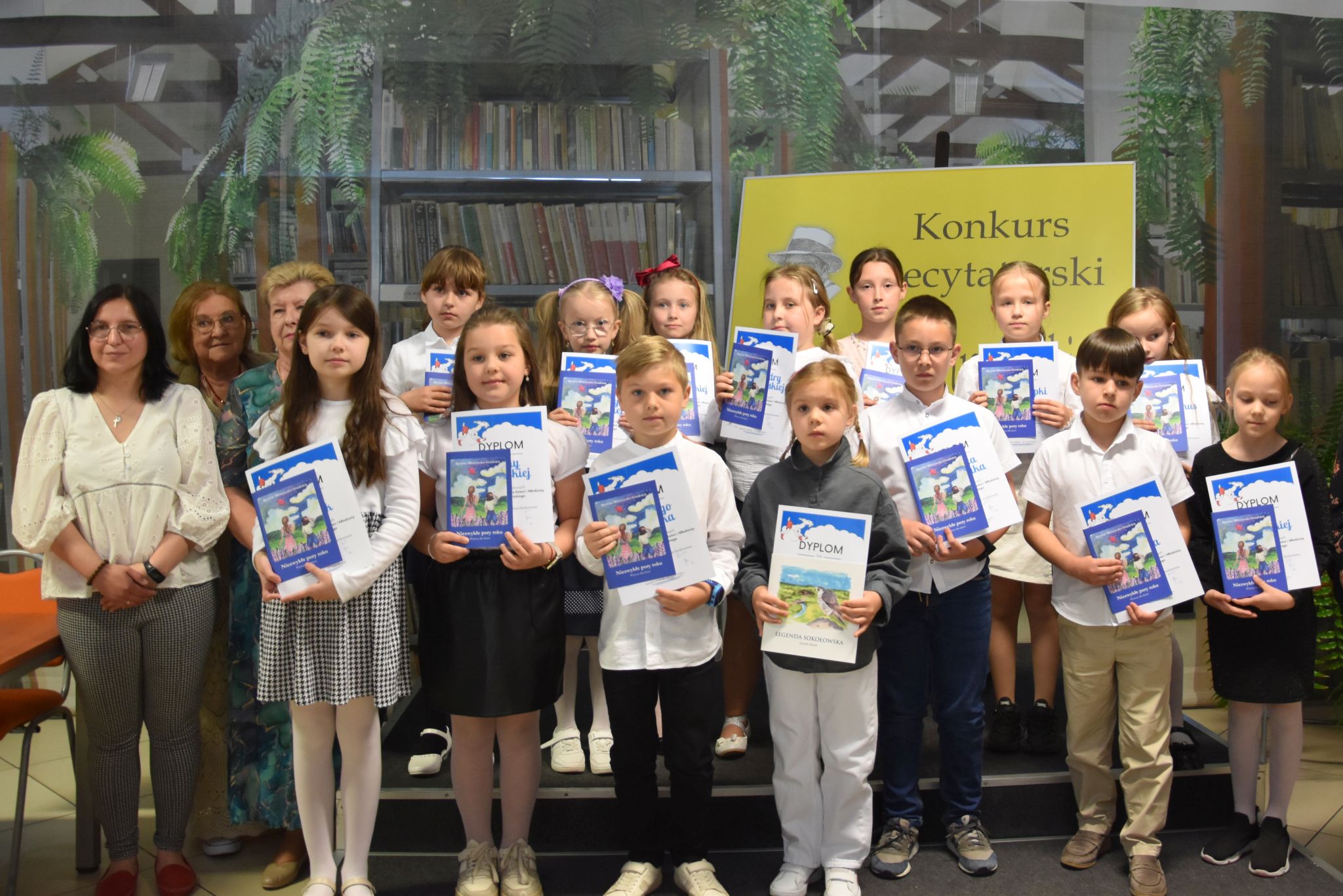 Grupa dzieci trzyma w rękach nagrody książkowe oraz dyplomy. Obok po prawej i lewej stronie stoją cztery kobiety. Trzy z komisji konkursowej oraz dyrektor biblioteki.