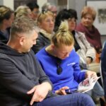 Uczestnicy Festiwalu Literatury Kulinarnej w sokołowskiej Bibliotece siedzą na widowni. Kobieta w niebieskiej bluzie z napisem 'Los Angeles' przegląda broszurę, obok niej mężczyzna w czarnej bluzie. W tle widać inne osoby zasłuchane w przebieg spotkania.