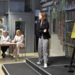 Młoda uczestniczka w czarnej marynarce i szarych spodniach stoi na scenie recytując utwór przed stołem z trzema członkiniami jury oraz żółtym banerem „Konkurs Recytatorski im. Kornela Makuszyńskiego” w bibliotece.