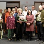 Czternaście osób stoi na małej scenie w bibliotece, po środku Lucyna Aleksandrowicz-Pędich trzyma bukiet różowych kwiatów, obok niej dyrektor biblioteki Małgorzata Iwańska-Kania.