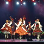 Na scenie o ciemnym tle znajduje się osiem osób – kobiety w haftowanych spódnicach z tiulowymi halkami, koralikami i kwiatowymi wiankami oraz mężczyźni w granatowych kaftanach z czerwonymi pasami i czapkach z piórami. Tancerze tworzą półokrąg i w dynamicznym ruchu unoszą spódnice, prezentując tradycyjny polski taniec folklorystyczny. Nad nimi wisi rząd czerwonych i białych świateł scenicznych.