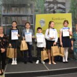 Grupa dzieci trzyma w rękach nagrody książkowe oraz dyplomy. Obok po prawej i lewej stronie stoją cztery kobiety. Trzy z komisji konkursowej oraz dyrektor biblioteki.