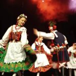 Na pierwszym planie młoda kobieta w białej bluzce z haftowanym gorsecikiem, zielono-czerwoną spódnicą i kwiatowym wiankiem na głowie unosi ramię, unosząc spódnicę. Obok niej mężczyzna w ciemnym kaftanie z czerwonymi wykończeniami i czapce z piórem wykonuje taniec, jego ruch jest lekko rozmyty. W tle widoczne kolejne postacie tancerzy, scena o ciemnym tle pod intensywnymi, pomarańczowo-białymi światłami sceny i delikatną mgiełką dymu.