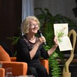 Lucyna Aleksandrowicz-Pędich podczas spotkania w bibliotece. Siedzi na pomarańczowym fotelu, trzyma w lewej ręce arkusz z akwarelową ilustracją kwiatów oraz zapisanym tekstem i mówi do mikrofonu trzymanego w prawej dłoni.