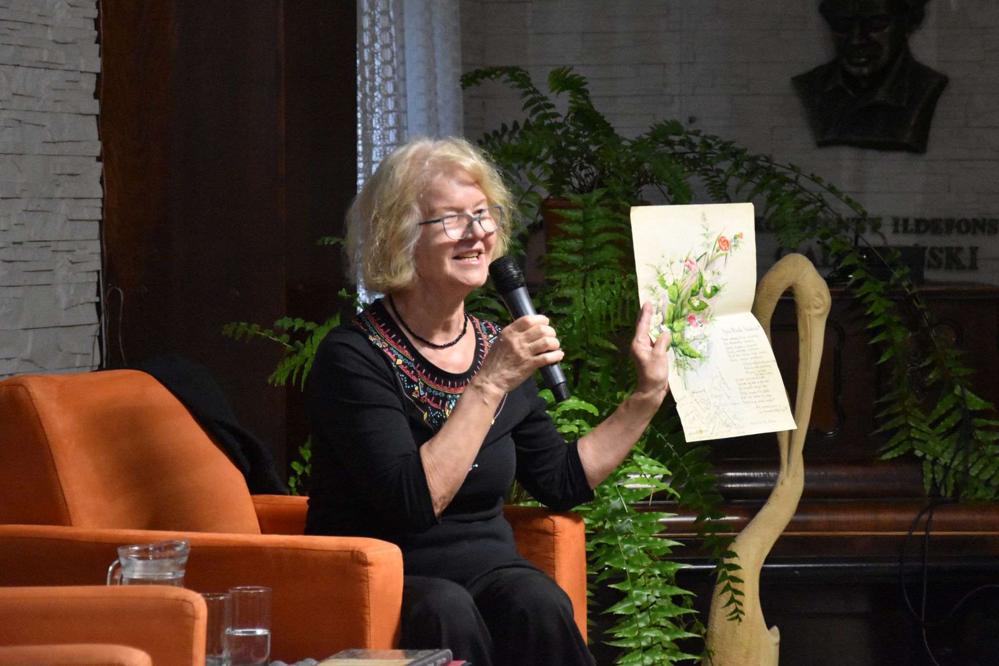 Lucyna Aleksandrowicz-Pędich podczas spotkania w bibliotece. Siedzi na pomarańczowym fotelu, trzyma w lewej ręce arkusz z akwarelową ilustracją kwiatów oraz zapisanym tekstem i mówi do mikrofonu trzymanego w prawej dłoni.