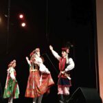 Na pierwszym planie tancerka w haftowanej sukience w czerwono-zielone wzory unosi spódnicę, a tancerz w granatowo-czerwonym kaftanie podnosi ramię, jakby zapraszając do kroku. Za nimi częściowo widoczna kolejna kobieta w zielonej spódnicy. Scena ma ciemne tło, nad głowami tancerzy wiszą reflektory emitujące ciepłe światło i delikatną mgiełkę dymu scenicznego.