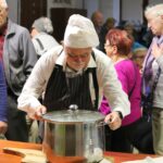 Pan Ryszard Szczepan Przytulski w kucharskim czepku i fartuchu ustawia garnek z zalewajką podczas Festiwalu Literatury Kulinarnej w sokołowskiej Bibliotece. W tle uczestnicy wydarzenia czekają na degustację.”