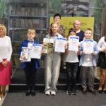 Grupa dzieci trzyma w rękach nagrody książkowe oraz dyplomy. Obok po prawej i lewej stronie stoją cztery kobiety. Trzy z komisji konkursowej oraz dyrektor biblioteki.