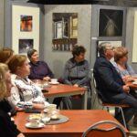 Na pierwszym planie grupa kilku osób siedząca przy stolikach podczas spotkania na holu w bibliotece. Za nimi na ścianie wiszą obrazy i fotografie związane z tematyką wiejską. Jedna z fotografii przedstawią drewniany wiatrak oświetlony nocą.