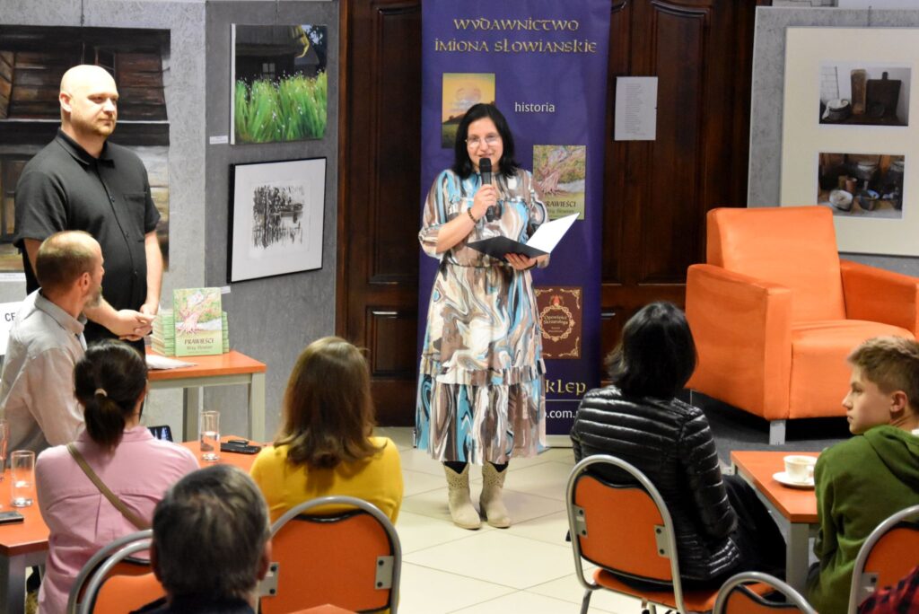 Na holu stoi dyrektor biblioteki Małgorzata Iwańska-Kania podczas przywitania autora Bartłomieja Dejnega. Za nimi znajduje się wystawa obrazów.