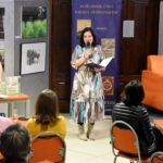 Na holu stoi dyrektor biblioteki Małgorzata Iwańska-Kania podczas przywitania autora Bartłomieja Dejnega. Za nimi znajduje się wystawa obrazów.